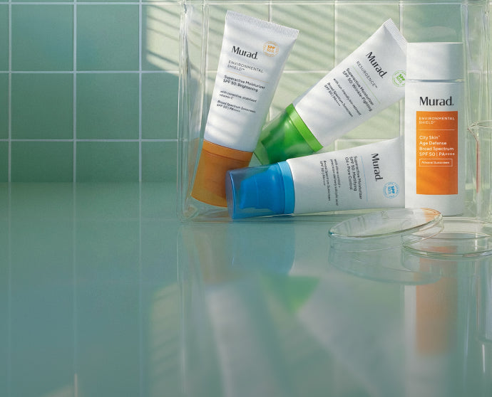 Four Murad skincare tubes and a bottle arranged in a clear plastic bag on a glossy teal surface, with a tiled wall and soft window-light shadows in the background.